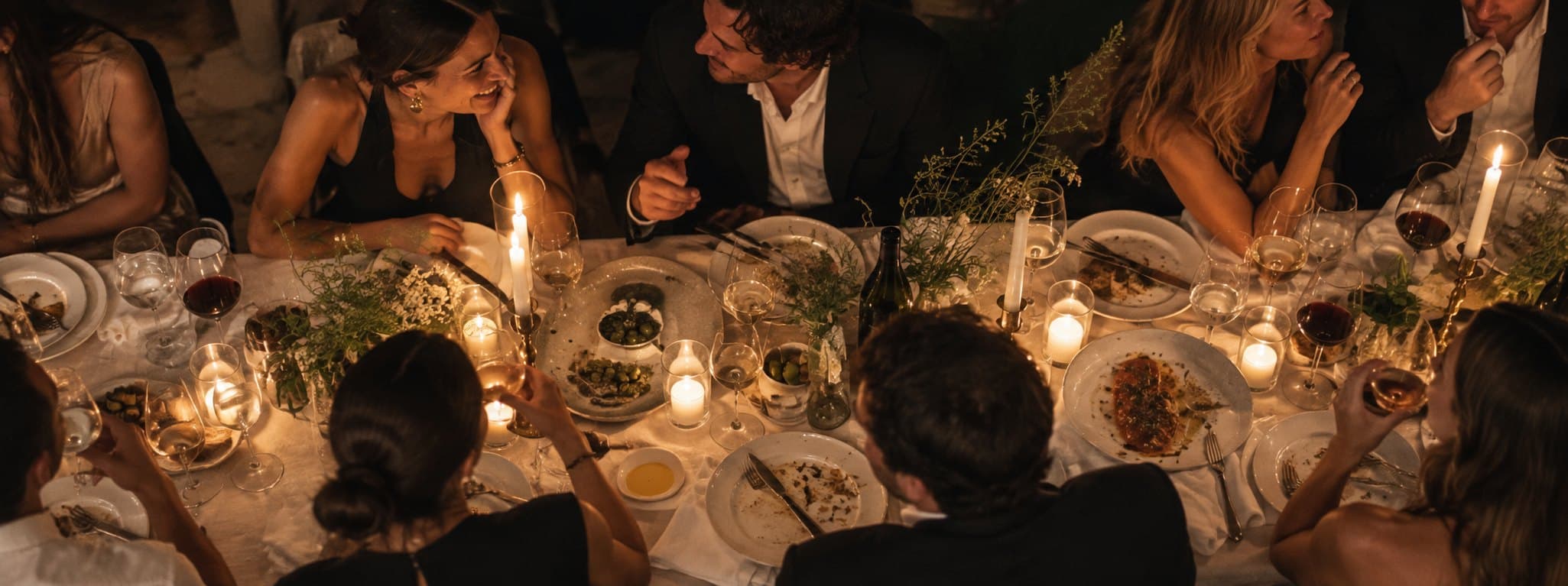 Guests around a candlelit dinner table seen from above, mid-conversation